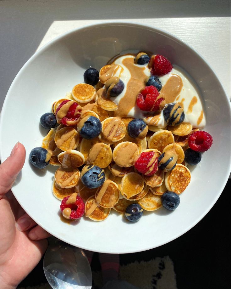 a person holding a white plate with pancakes and berries on it next to a spoon