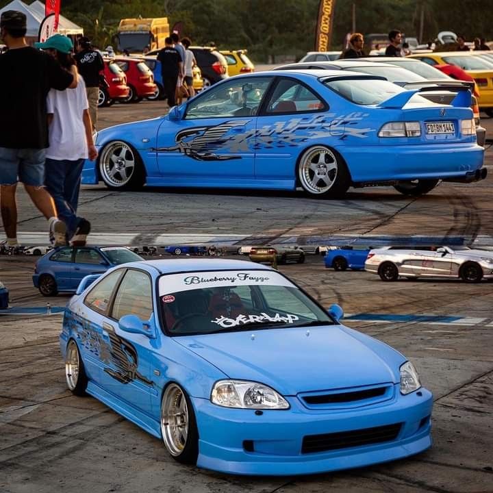 two blue cars parked next to each other in a parking lot with people looking at them