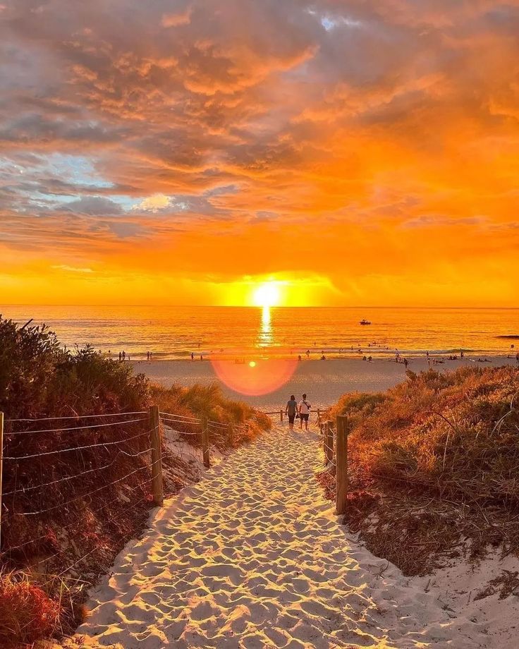 the sun is setting over the beach and people are walking down the path to the water