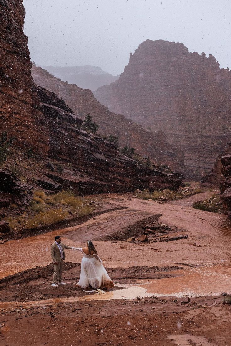 a man and woman standing in the middle of a dirt road with mountains in the background