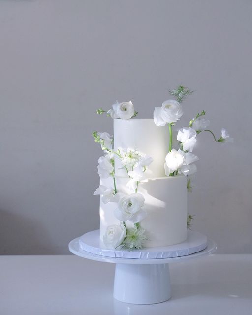 a three tiered cake with white flowers and greenery on the top is sitting on a pedestal