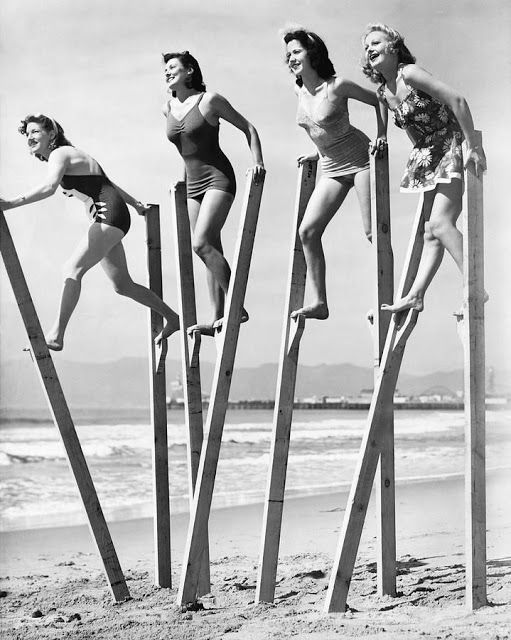 four women in bathing suits standing on stilts at the beach, all leaning forward