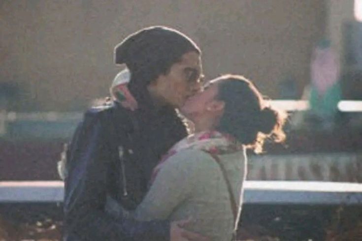 two people standing next to each other in front of a park bench and one is kissing the other