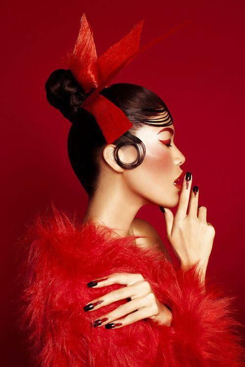 a woman with red hair and black nails wearing a red feathered dress, holding her hand to her face