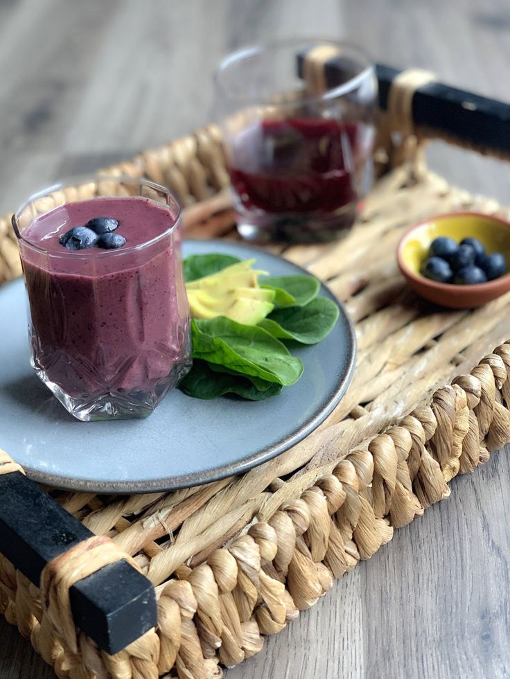 two glasses of blueberry smoothie on a wicker tray next to some fruit