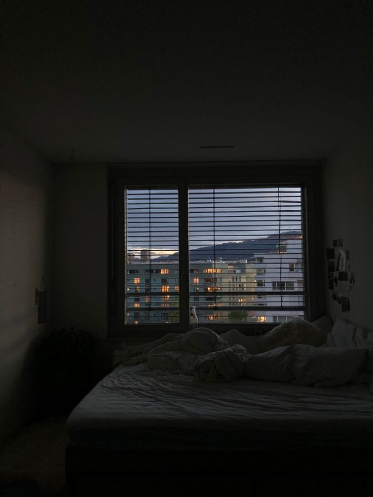 an unmade bed sitting in front of a window with the city lights seen through it