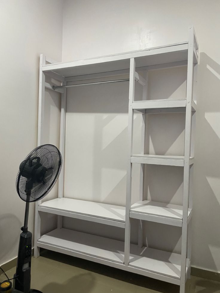 a room with shelves and a fan on the floor in front of it, all white