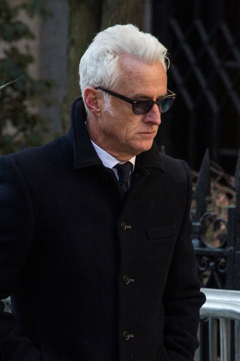 an older man with white hair and glasses is walking down the street wearing a black coat