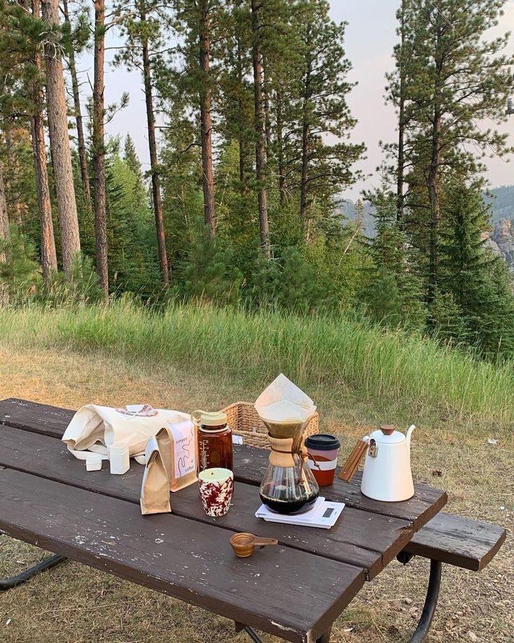 a picnic table with coffee and tea on it in the middle of a field surrounded by trees