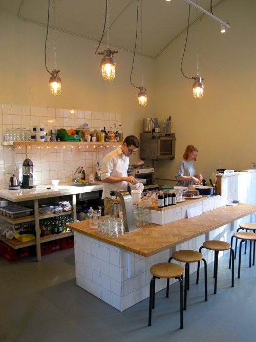 two people preparing food in a kitchen with lots of counter space and lights hanging from the ceiling