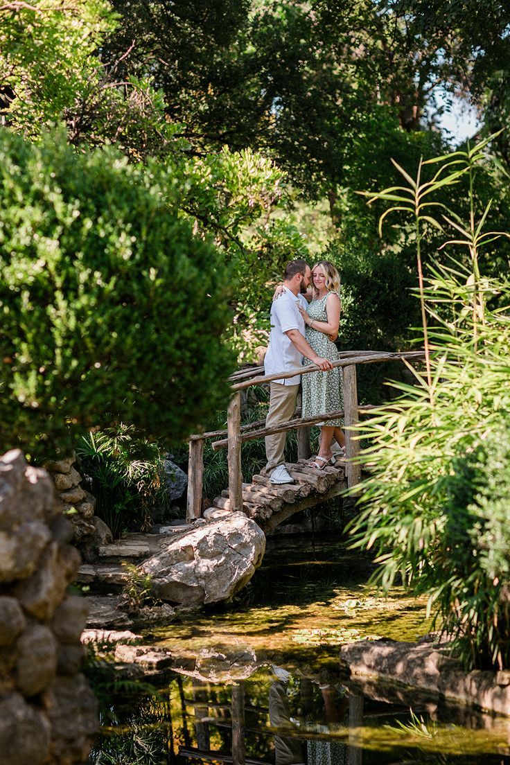 a man and woman standing on a bridge in the middle of a pond surrounded by trees