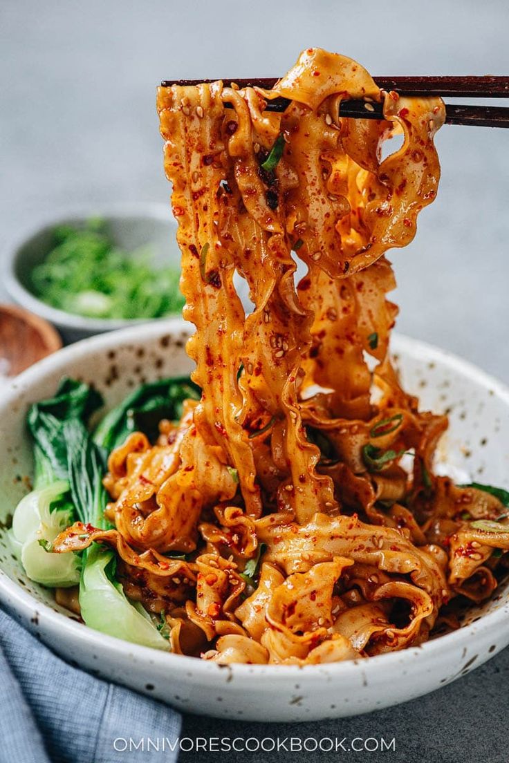 chopsticks sticking out of a bowl of noodles with vegetables and sauce on the side