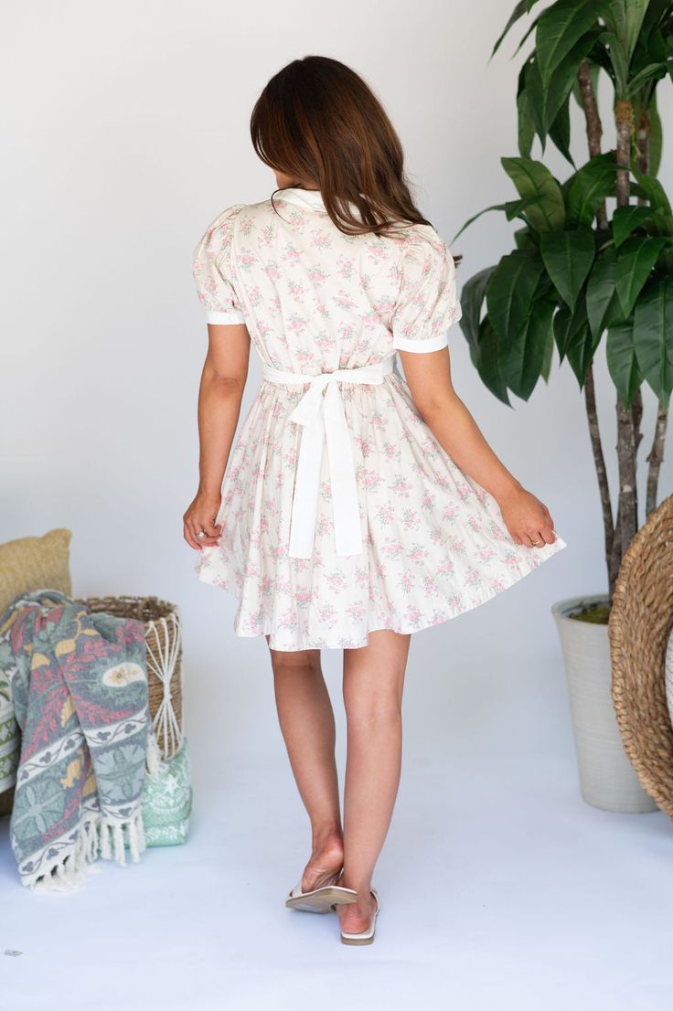 Expertly designed Claire Collar Mini Dress boasts a pink and green floral print, contrasting with a crisp white collar and quarter pearl button closures along the front. Short puff sleeves and a white waist tie add feminine charm, while a side zipper closure ensures a secure fit! 100% rayon, lined Model is 5'3" with a 32" bust, 25" waist, and 38" hips and wearing a small. Size recs: 0-2:S, 4-6:M, 8:L White Collared Dress With Tie Waist, White Collared Dress For Garden Party, White Peter Pan Collar Dress For Garden Party, Spring Cream Dress With Peter Pan Collar, Cream Collared Spring Dress, Collared Cream Spring Dress, Collared Cream Dresses For Spring, Cream Short Sleeve Floral Dress For Garden Party, Cream Floral Short Sleeve Dress For Garden Party