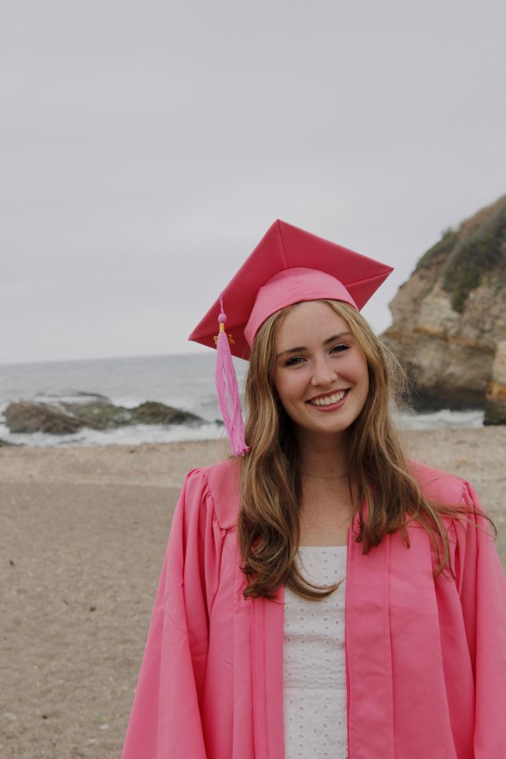 a young woman wearing a pink graduation cap and gown stands on the beach in front of the ocean