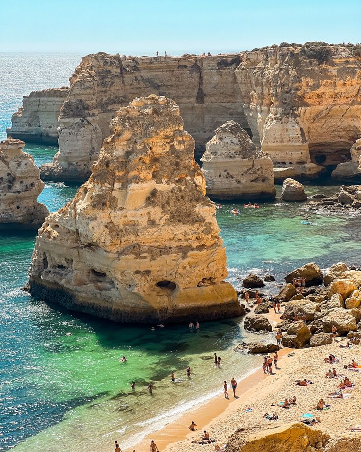 many people are swimming in the water near some rocks and cliffs on a beach with clear blue water