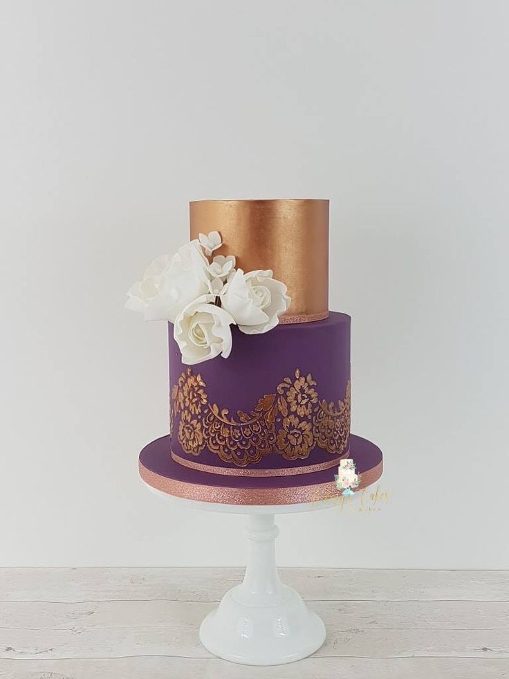 a purple and gold wedding cake with white flowers on the top, sitting on a pedestal