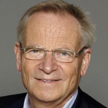 an older man wearing glasses and a suit smiling at the camera with a gray background