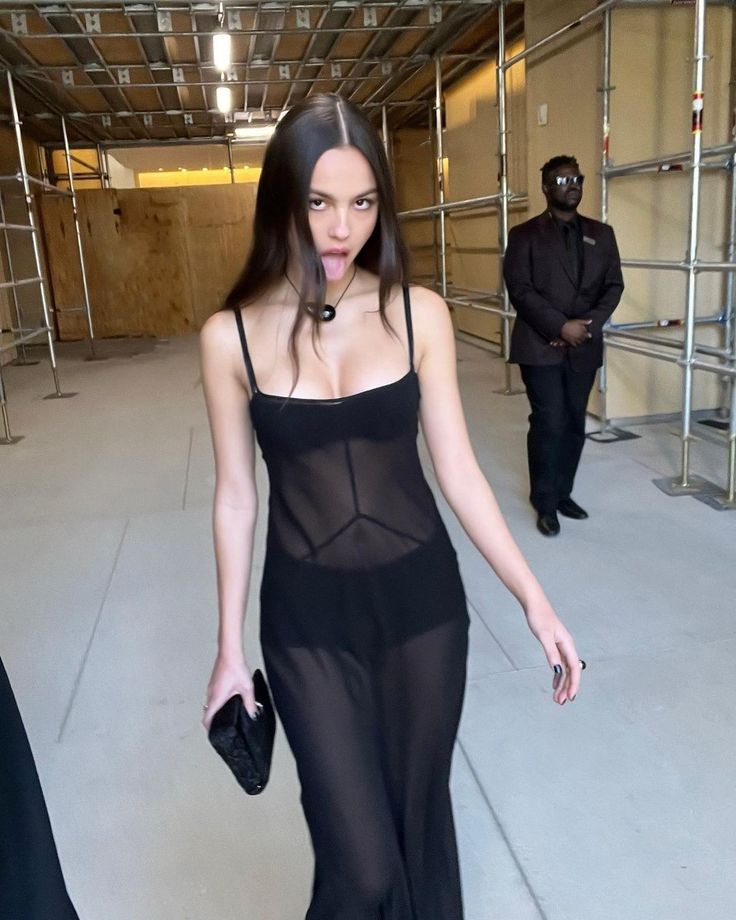 a woman walking down a runway wearing a black dress and holding a handbag in her right hand