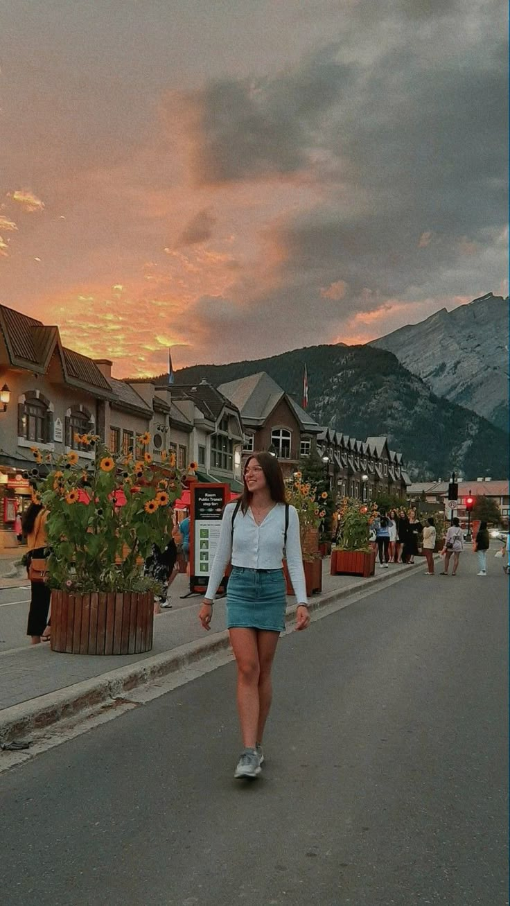 a woman is walking down the street in front of some buildings and flowers at sunset