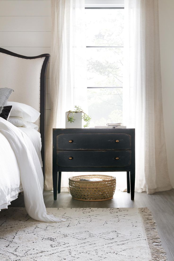 a bedroom with a white bed and black dresser in front of the window, along with a basket on the floor