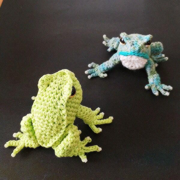 two crocheted frogs sitting next to each other on a black surface with one frog looking at the camera