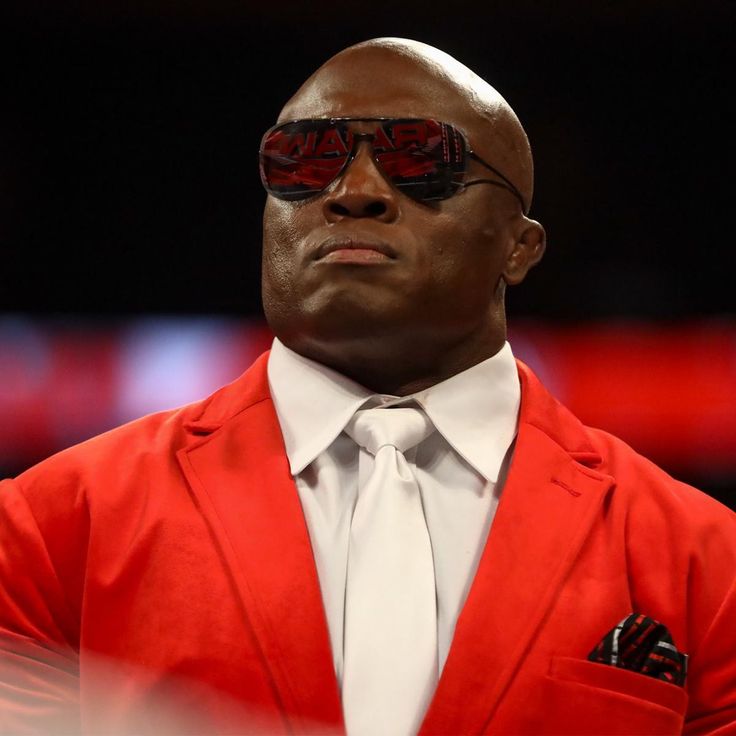 the rock wearing sunglasses and a red suit looks off into the distance while standing in front of an audience