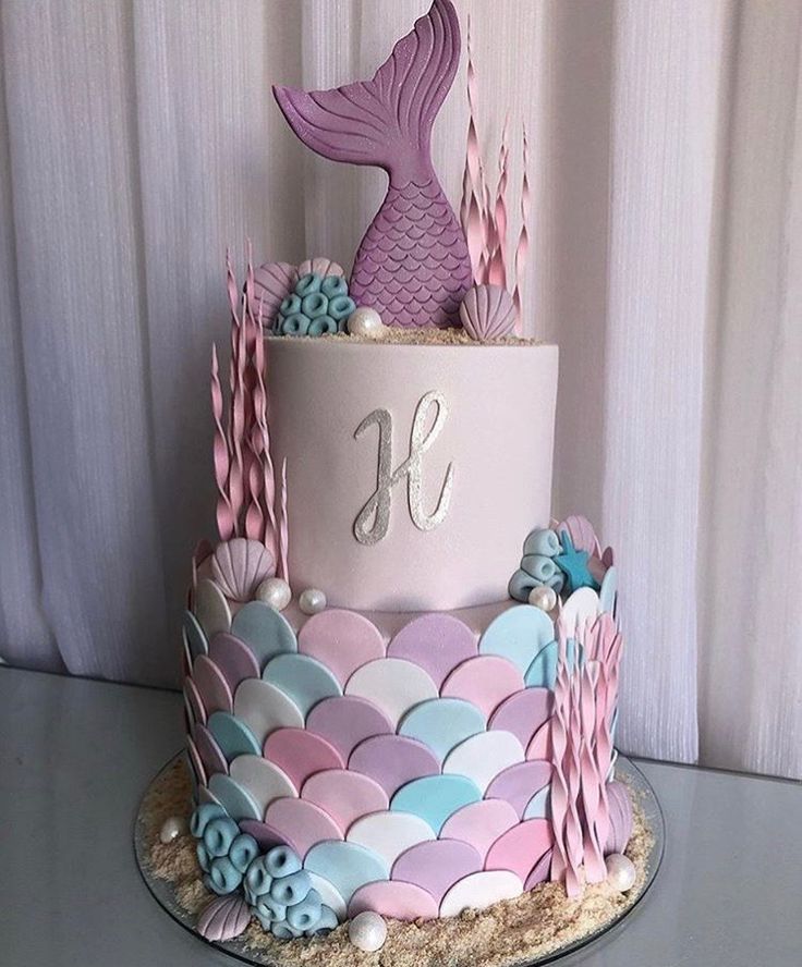 a three tiered cake decorated with pink, blue and white mermaid tail decorations on a silver platter