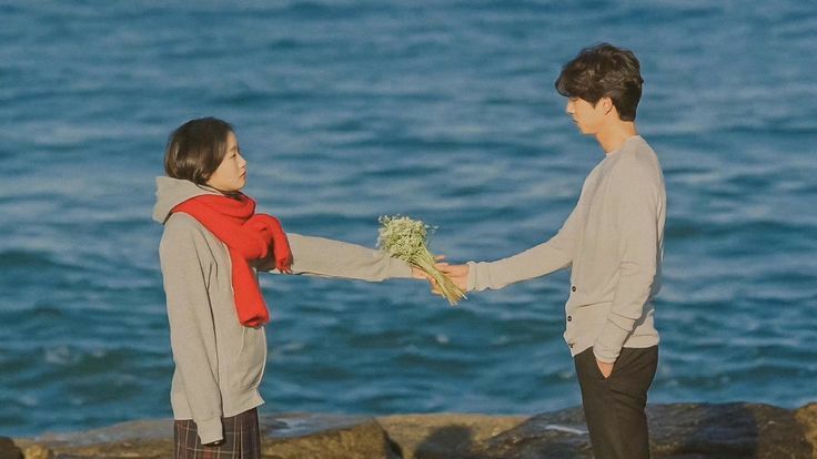 two people standing on rocks near the water holding each other's hands and looking at each other