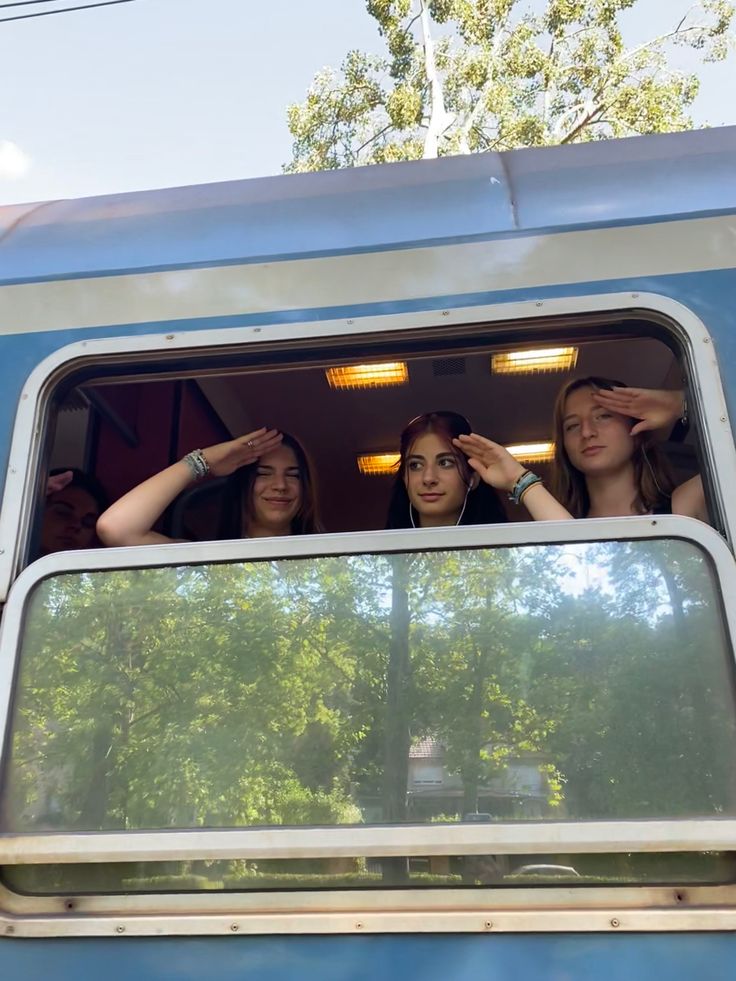 three girls are looking out the window of a blue train with trees in the background