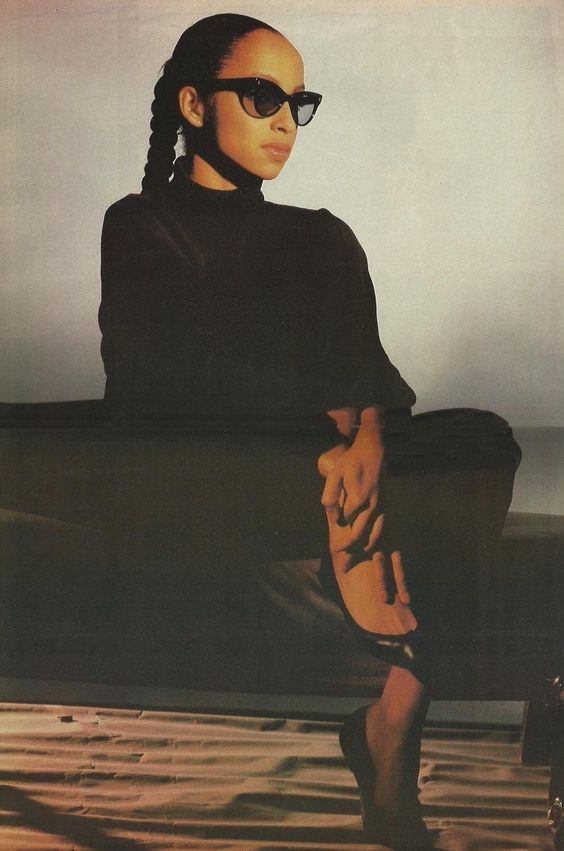 a woman sitting on top of a black couch wearing sunglasses and a turtle neck sweater