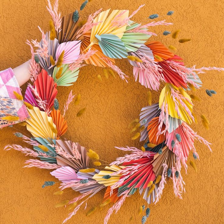 a colorful wreath made out of folded paper on a yellow wall with hands holding it