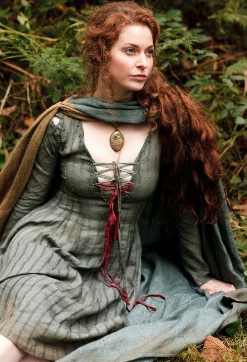 a woman with long red hair sitting on the ground wearing a green dress and cloak