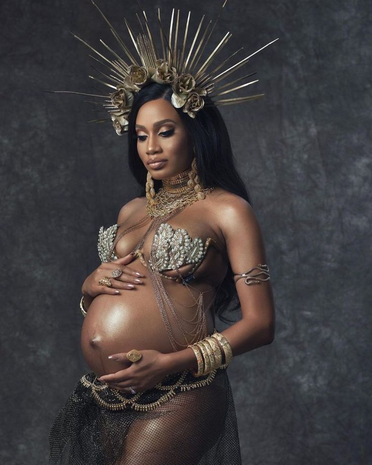 a pregnant woman wearing gold jewelry and holding her belly in front of a dark background