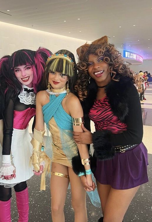 three women dressed up in costumes posing for the camera at an airport terminal, with one woman holding her arm around the other's waist