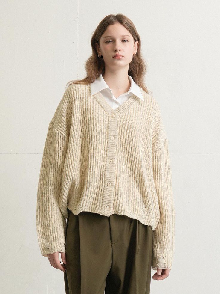 a woman standing in front of a white wall wearing a beige sweater and khaki pants