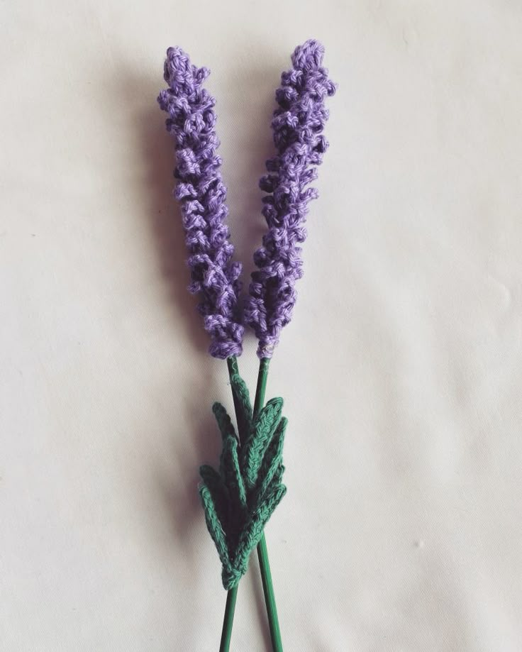 two purple flowers with green stems on a white tableclothed surface, one is crocheted and the other is crocheted