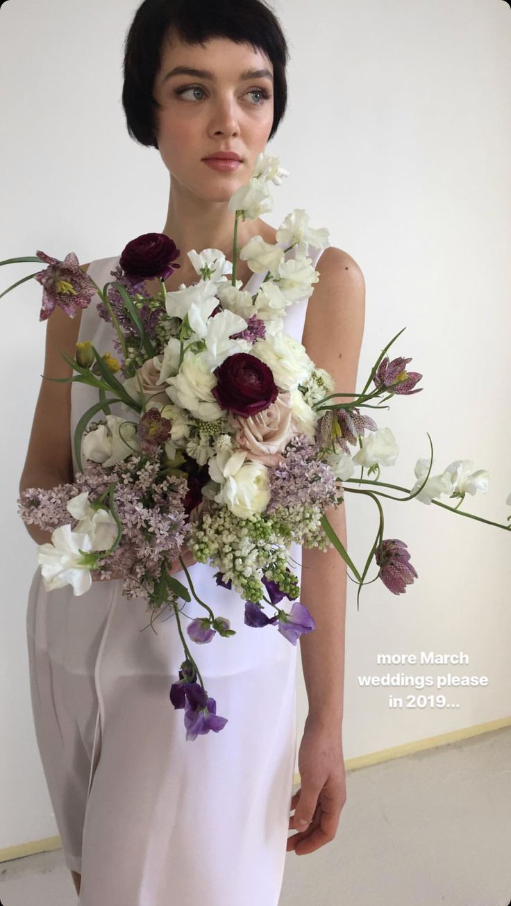 a woman holding a bouquet of white and purple flowers in front of her face while standing next to a wall