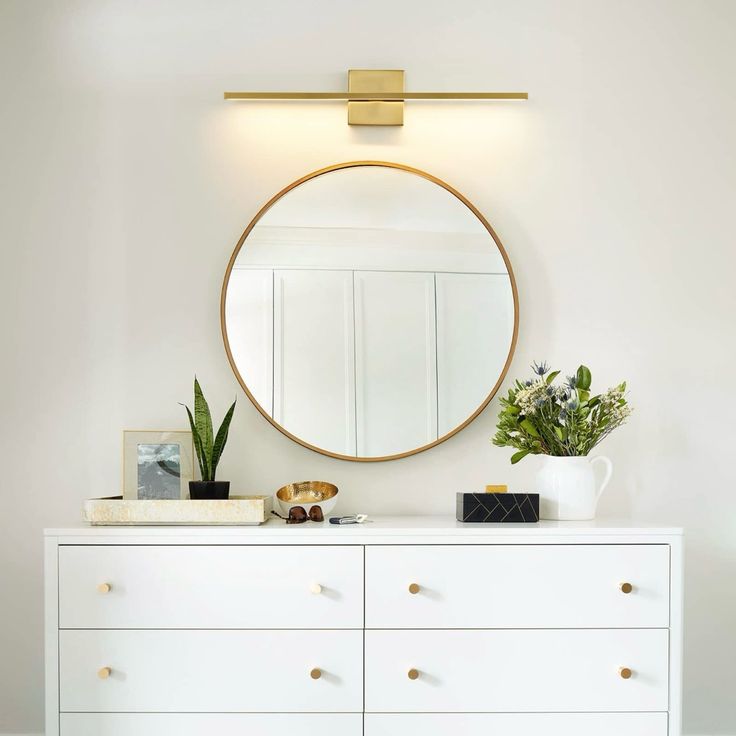 a white dresser topped with a round mirror and potted plant next to a gold framed mirror