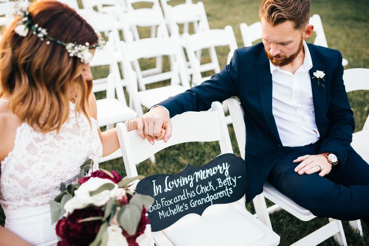 a bride and groom sitting on white chairs holding hands