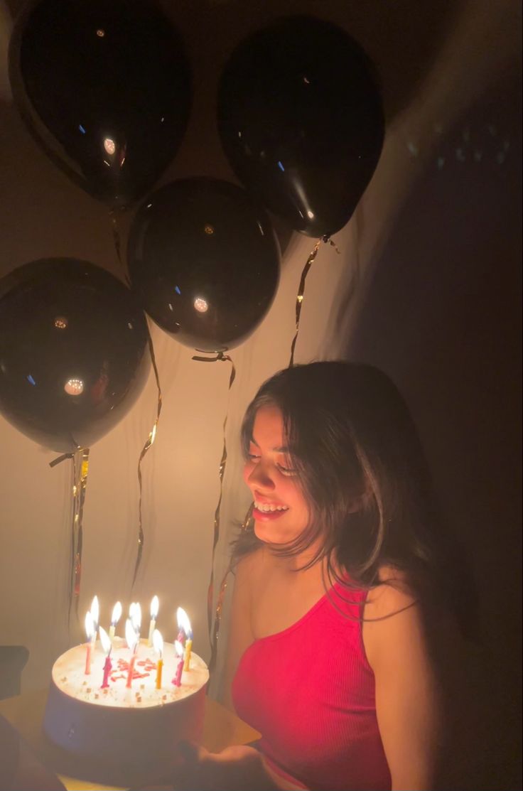 a woman holding a birthday cake with lit candles in front of her, surrounded by balloons