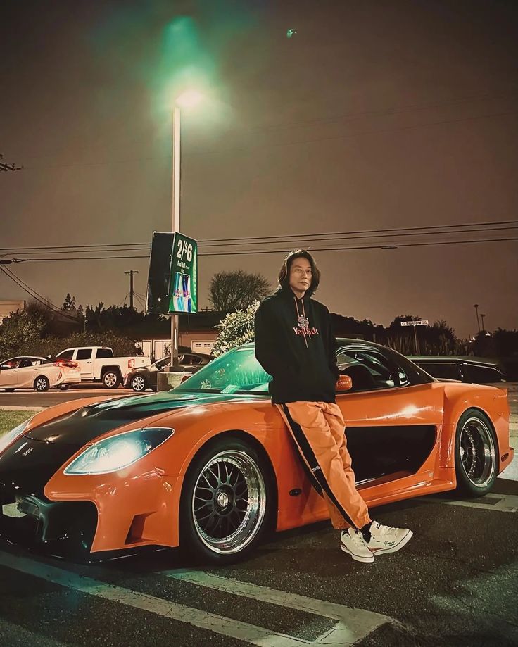 a man sitting on top of an orange sports car in front of a gas station