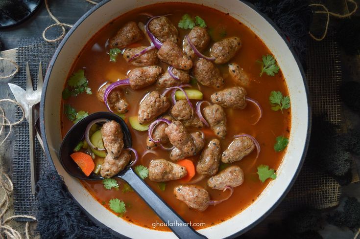 a bowl filled with meatballs, carrots and onions next to a spoon on a table