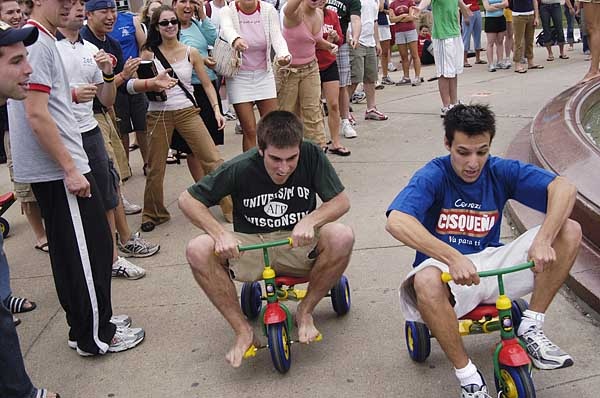 two young men riding tricycles in front of a fountain with people standing around