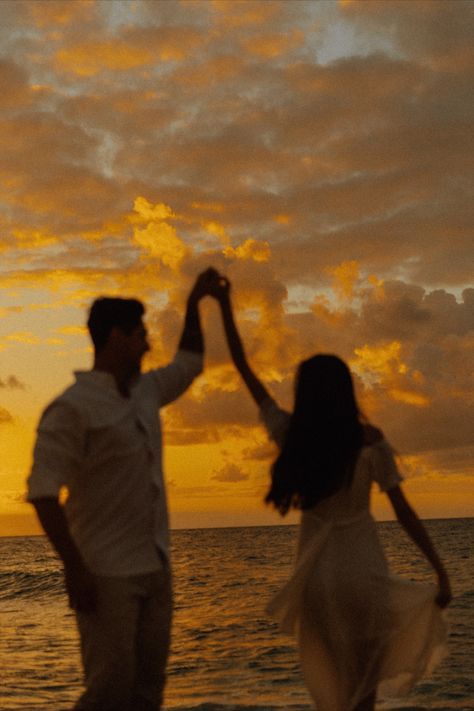 a man and woman holding hands while walking on the beach at sunset with clouds in the background