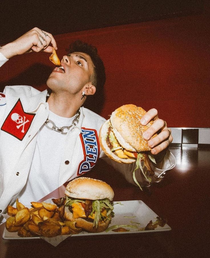 a man in white jacket eating a large hamburger next to french fries and potato wedges