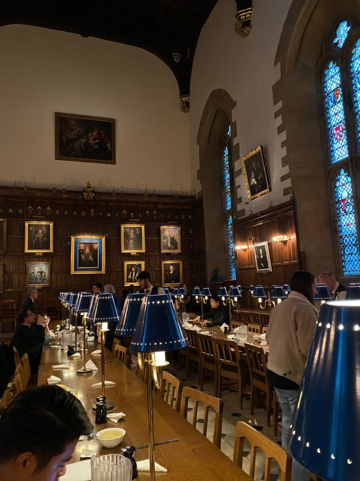 people sitting at tables in a large dining hall with stained glass windows and blue lamps