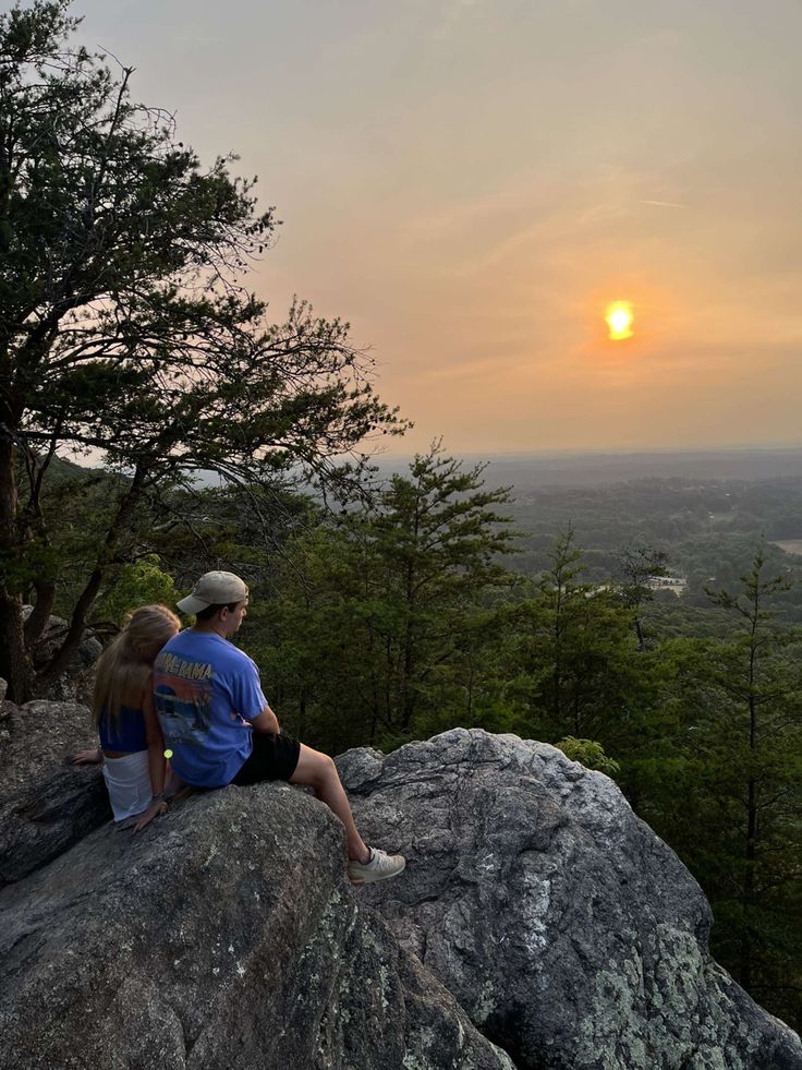 two people sitting on top of a large rock next to a forest at sunset with the sun in the distance