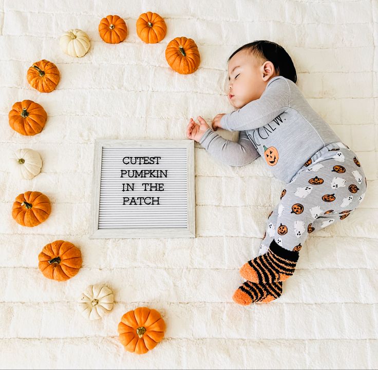 a baby laying on top of a bed next to pumpkins and a sign that says cutest pumpkin in the patch