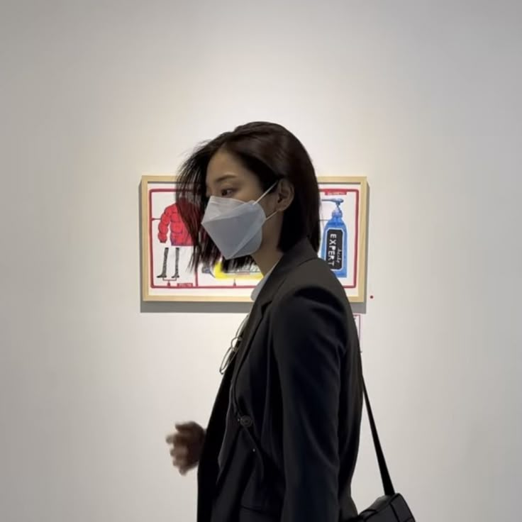 a woman wearing a face mask in front of a wall with pictures on it and a handbag