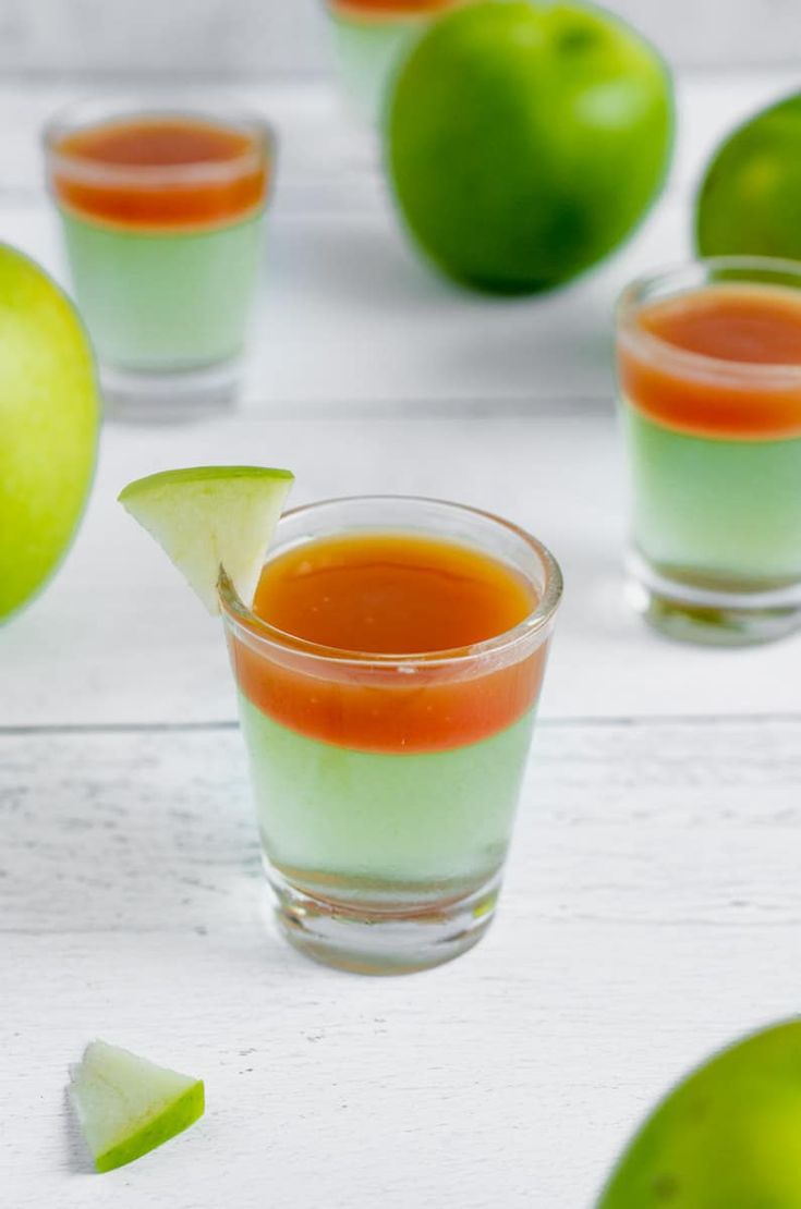 small glasses filled with green and orange jello sitting next to apples on a white table